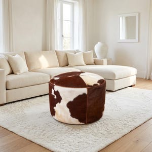 May include: A round ottoman upholstered in genuine cowhide featuring a natural pattern of dark brown and cream patches. The cylindrical piece has visible stitched seams and a smooth, low-profile design, providing a textured focal point.