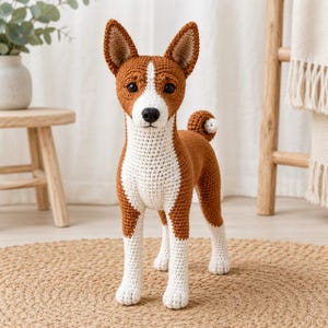 Puede incluir: Una figura de perro tejida a ganchillo hecha a mano con pelaje marrón y blanco. El perro tiene orejas puntiagudas erguidas, nariz negra y cola enroscada. La construcción de hilo muestra un patrón de puntadas detallado en todo el cuerpo.