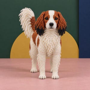 以下が含まれることがあります： 白、茶、黒の配色で手作りされたかぎ針編みの犬のフィギュア。このぬいぐるみは、質感のある毛並み、黒い先端を持つ長い垂れ耳、ふさふさした直立した尻尾、そして暗い目をしており、リアルな立ち姿をしています。
