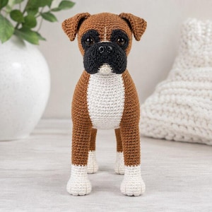 May include: A crocheted boxer dog figurine featuring a brown body, a black muzzle, and a white chest and paws. The toy has dark brown eyes and a black nose, with a textured yarn surface created through detailed stitching throughout the entire body.