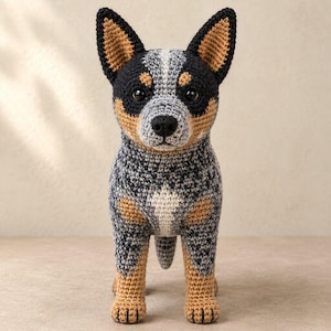 May include: A crocheted Australian Cattle Dog figurine featuring a speckled gray, black, and white coat with tan markings on the ears, face, and legs. The plush toy has upright pointed ears, dark plastic eyes, and a black nose, standing upright.