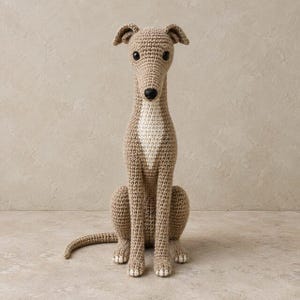 May include: A crocheted stuffed animal in the shape of a greyhound dog. The figure is light brown with a white patch on its chest, black plastic eyes, and a black nose. It is depicted in a seated position with its tail extended to the side.