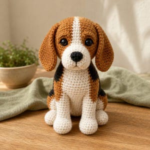 Op de afbeelding: Een gehaakt beagle-hondfiguurtje met een driekleurig patroon van bruin, zwart en wit garen. Het knuffeldier heeft grote, donkere, ronde ogen, hangende oren en een kleine zwarte neus, gemaakt met een zichtbare textuursteek over het geheel.
