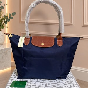 May include: A navy blue nylon tote bag featuring a brown leather flap with a gold-tone snap button closure. The bag has two handles wrapped in protective white paper and a white Longchamp brand tag attached to the side with a green string.