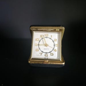 May include: A vintage Equity travel alarm clock featuring a square white face with gold-toned numerals and hands. The clock is housed in a black, hinged case with gold-colored metal trim, designed to fold open for display on a flat surface.