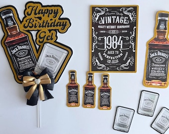 May include: A set of Jack Daniel's themed cake toppers including a Happy Birthday Gel sign, a large bottle cutout, a rectangular vintage 1984 sign, three small bottle cutouts, three glass cutouts, and a black and gold ribbon bow on a stick.