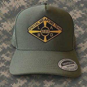 Può includere: Un cappellino da baseball verde oliva con chiusura snapback e un adesivo circolare sulla visiera. La parte anteriore presenta una toppa a forma di diamante con testo giallo SPECIAL WEAPONS TEAM e SEB, con le lettere L, A, S, D negli angoli.