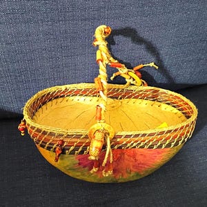 Pode incluir: Uma cesta de formato oval com interior amarelo claro e exterior pintado com padrões florais rosa e verdes. A borda é trançada com detalhes em marrom escuro e vermelho, e uma alça única é decorada com várias contas de madeira.