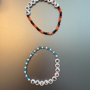 May include: Two beaded bracelets. The top bracelet features white letter beads spelling POOH PAVEL with a red heart bead, followed by orange, red, and black seed beads. The bottom bracelet features white letter beads spelling SAILUB PON with a red heart bead, followed by blue and clear seed beads.