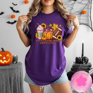 Peut inclure: Un t-shirt violet avec une impression graphique représentant une tasse de café, une citrouille avec un nœud rose, un ballon de football américain, des tournesols et des feuilles d'automne. Le texte "JUST A GIRL WHO LOVES FALL" est imprimé en orange.