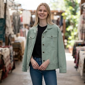 May include: A light sage green denim jacket featuring a classic collar, long sleeves, and a front button closure. The jacket includes two chest pockets with buttoned flaps and is worn over a plain black shirt and blue denim jeans.