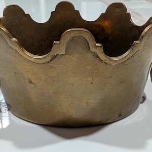 Puede incluir: Un cuenco de latón con borde festoneado y dos asas en forma de cabeza de león a los lados. El cuenco tiene un acabado metálico en tono dorado y una base redondeada y lisa. Las cabezas de león tienen pequeños anillos circulares colgando.