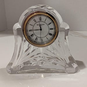 May include: A clear crystal mantel clock with a faceted, arched design. The round clock face features a gold-tone bezel, black Roman numerals, and the text Waterford Crystal printed in black at the center with black hour, minute, and second hands.