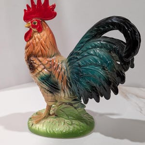 May include: A ceramic rooster figurine featuring a vibrant red comb and wattle, a golden-brown neck, and a glossy tail with deep blue and black feathers. The bird stands on a textured green base, showcasing detailed plumage and a realistic posture.