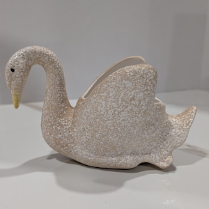 May include: A ceramic swan-shaped planter or decorative bowl featuring a textured, speckled beige and white finish. The swan has a long, curved neck, a small black eye, and a yellow beak, with an open cavity on its back for holding items.