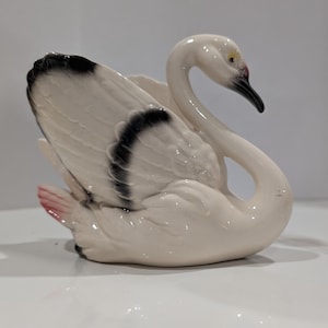 May include: A white ceramic swan figurine with a curved neck and black-tipped wings. The swan features a glossy finish, a black beak with a hint of pink at the base, and subtle pink coloring on the tail feathers, presented in a graceful pose.