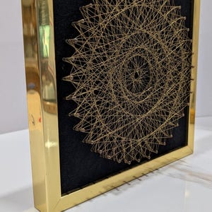 May include: A square wall art piece featuring a circular string art design made of thin gold thread wrapped around small nails. The design is set against a black background and enclosed within a polished, reflective gold-colored metal frame.