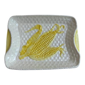May include: A rectangular ceramic serving platter featuring a cream-colored basket-weave texture across the surface. The center displays a raised, three-dimensional relief of two yellow corn cobs with husks. The edges have a rope-like border design.
