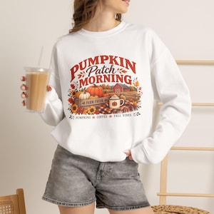 Peut inclure: Un sweat-shirt blanc à manches longues avec une impression graphique portant le texte PUMPKIN PATCH MORNING. Le motif représente une caisse en bois de citrouilles, une grange rouge, des tournesols, une couverture à carreaux et une tasse.