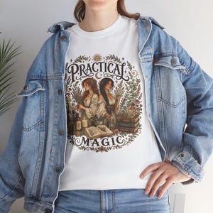 May include: A white t-shirt featuring a colourful graphic print of two figures surrounded by botanical elements, books, and candles. The text Practical Magic is arched above and below the illustration, which is framed by a circular floral wreath.