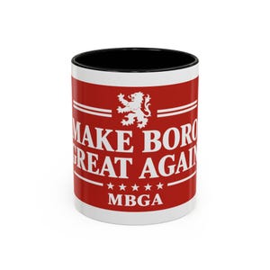 May include: A white ceramic mug with a black interior and rim. The exterior features a red band with white text reading MAKE BORO GREAT AGAIN, a white lion emblem above the text, five white stars below, and the acronym MBGA at the bottom.