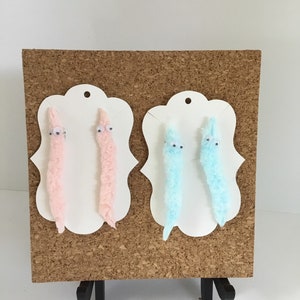 Puede incluir: Dos pares de pendientes de gusano peludos, un par es rosa y el otro es azul. Los pendientes se muestran en un tablero de corcho con cartulina blanca detrás.