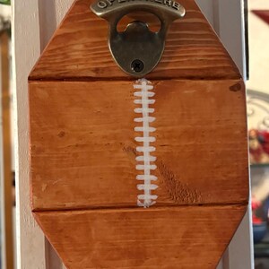 May include: A wall-mounted bottle opener shaped like an American football, crafted from light brown wood with a vertical white painted lace pattern. A metallic bronze-toned opener is attached at the top with the embossed text OPEN HERE.