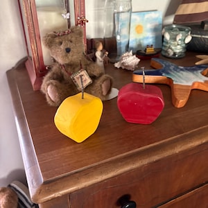 May include: Two solid, apple-shaped wooden blocks, one painted bright yellow and the other painted deep red, each featuring a single metal nail protruding vertically from the top center, designed to function as a memo or note holder.