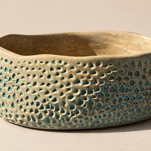 May include: A round ceramic bowl with an uneven, wavy rim. The exterior features a textured surface with numerous small, indented dimples that reveal a bright turquoise blue glaze within the depressions against a matte beige clay finish.