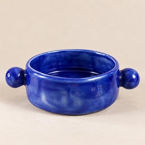 Peut inclure: Un bol rond en céramique avec un vernis bleu profond et une finition lisse et brillante. Le bol présente deux poignées sphériques sur les côtés opposés, s'étendant vers l'extérieur depuis le bord. Les surfaces montrent des nuances de bleu.