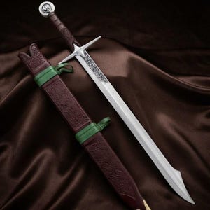 Peut inclure: Une épée en métal argenté avec une poignée brune texturée et un pommeau circulaire. La lame présente des gravures complexes près de la garde. À côté se trouve un fourreau brun texturé assorti avec des bandes vertes et une pointe en métal doré.