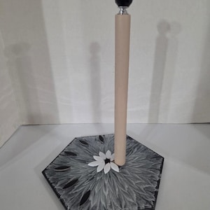 May include: A paper towel holder featuring a hexagonal base with a gray and white textured pattern, accented by black petal-shaped inlays and a central white flower design. A vertical wooden dowel stands in the center, topped with a black faceted finial.
