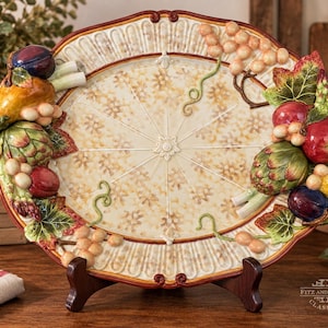 May include: An oval ceramic serving platter featuring a cream-colored center with a subtle floral pattern and a raised, hand-painted border decorated with three-dimensional fruits and vegetables, including grapes, artichokes, and pears.