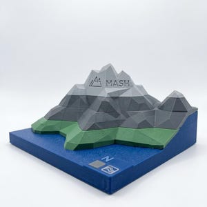 May include: A 3D printed low-poly mountain model featuring a grey peak with the text MASH, a green base representing land, and a blue base representing water. The blue base includes a small square with the letter N and a wave icon symbol.
