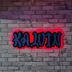 May include: A custom wall sign featuring the name KALVIN in bold, blue, graffiti-style lettering. The sign is mounted on a black backing and illuminated by a bright red LED light that creates a glowing halo effect against the wall surface.