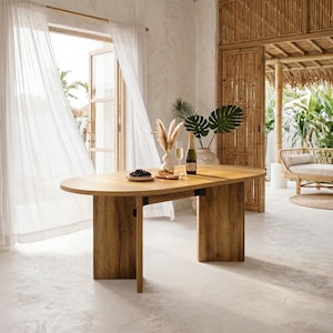May include: An oval wooden dining table with a light brown finish and two wide, flat vertical supports. The tabletop holds a small plate of dark grapes, a glass of white wine, a bottle of champagne, and a white vase containing dried pampas grass.