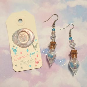 May include: A pair of drop earrings featuring miniature glass bottles filled with small blue, white, and iridescent beads, topped with cork stoppers and clear crystal accents. Beside them is a cream-coloured tag with the text Trans-for-Magic.
