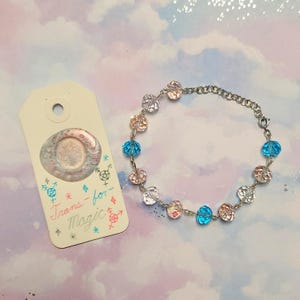 Peut inclure: Un bracelet composé de perles à facettes rose clair, blanches et bleu vif, reliées par une chaîne en métal argenté. À côté se trouve une étiquette rectangulaire avec un sceau de cire et le texte manuscrit Trans-for-Magic.
