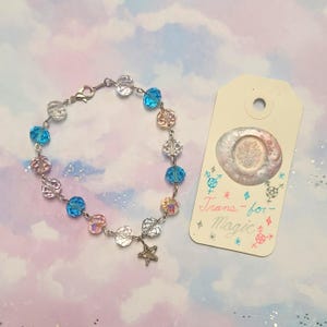 Peut inclure: Un bracelet composé de perles à facettes alternées transparentes, rose clair et bleu vif sur une chaîne argentée avec une petite breloque étoile, accompagné d'une étiquette rectangulaire avec un sceau de cire et le texte Trans-for-Magic.