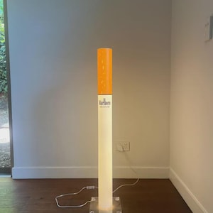 May include: A tall, cylindrical floor lamp designed to resemble a cigarette. The top section is orange, while the lower white section features the black text Marlboro MEDIUM. It stands on a clear square base and includes a white power cord.