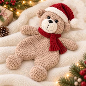 Pode incluir: Um urso de peluche em croché bege com um gorro de Pai Natal vermelho e branco e um cachecol vermelho a condizer. O urso tem olhos de plástico preto, um nariz bordado a preto e um focinho branco. O corpo é plano, macio e flexível.