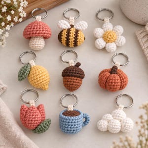 May include: A collection of nine crocheted keychains featuring various shapes including a pink mushroom, a yellow and black bee, a white daisy, a yellow lemon, a brown acorn, an orange pumpkin, a pink tulip, a blue coffee mug, and a white cloud.
