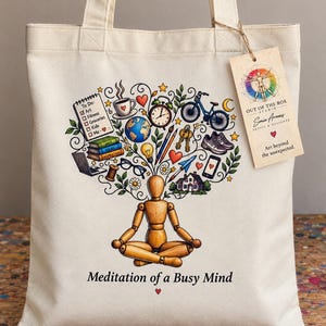 Peut inclure: Un sac fourre-tout en toile couleur crème représentant un mannequin en bois assis en position du lotus. Au-dessus, une illustration colorée montre divers objets dont une horloge, un vélo, un globe, des livres et une liste de tâches. Le texte "Meditation of a Busy Mind" est imprimé sous la figure et une étiquette de marque est attachée à la poignée.