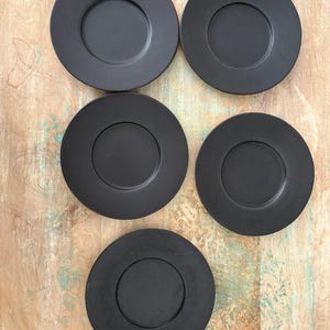 May include: A set of five circular black plates arranged in a cluster. Each plate features a smooth, matte finish with a distinct recessed center circle and a wide, flat rim. The plates are uniform in color and design, presenting a simple, functional aesthetic.