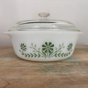 May include: A white milk glass casserole dish with a clear glass lid. The exterior of the bowl features a repeating green floral pattern consisting of stylised flowers and leafy stems. The dish has integrated side handles and a round glass knob.
