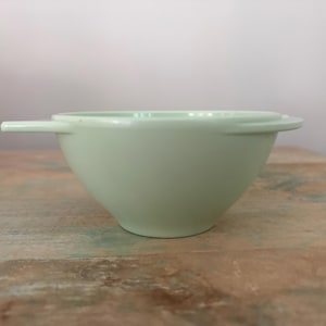 May include: A light mint green bowl with a smooth, matte finish. The bowl features a wide, flat rim and a single, thin, horizontal handle extending from one side. The design is simple and functional, with a rounded base and a wide opening.