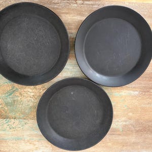 May include: Three circular, matte black plates arranged in a triangular formation. Each plate features a flat, wide base with a slightly raised rim, showcasing a dark, uniform finish with subtle surface textures visible across the flat centers.