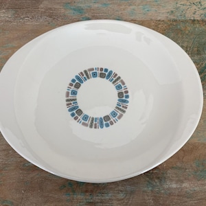 May include: A round white ceramic bowl featuring a central circular pattern composed of small, repeating geometric shapes in shades of blue, brown, and grey. The bowl has a smooth, glossy finish and a wide, shallow basin design.