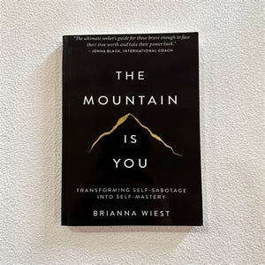May include: A black book titled The Mountain Is You by Brianna Wiest. The cover features a gold mountain graphic, the subtitle Transforming Self-Sabotage Into Self-Mastery, and a quote by Jenna Black at the top in white and gold text.