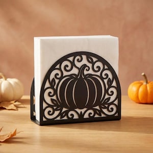 Puede incluir: Un servilletero de metal negro con diseño semicircular, que presenta una silueta de calabaza central rodeada de intrincados motivos de volutas y hojas, sosteniendo una pila de servilletas de papel cuadradas blancas en posición vertical.